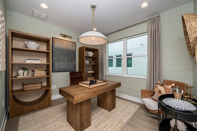 office area with wallpapered walls, light wood finished floors, baseboards, visible vents, and a textured ceiling