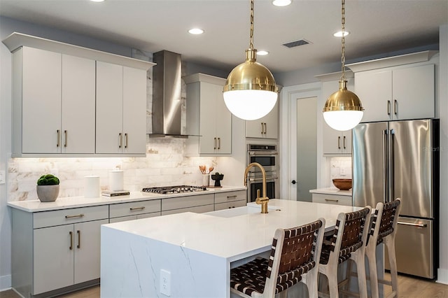 kitchen featuring appliances with stainless steel finishes, light countertops, wall chimney range hood, and an island with sink