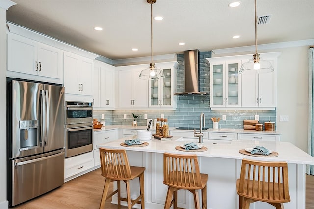 kitchen with stainless steel appliances, decorative light fixtures, a kitchen island with sink, and wall chimney range hood