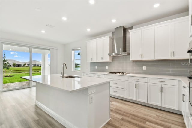 kitchen featuring gas cooktop, a kitchen island with sink, a sink, light wood-style floors, and wall chimney range hood