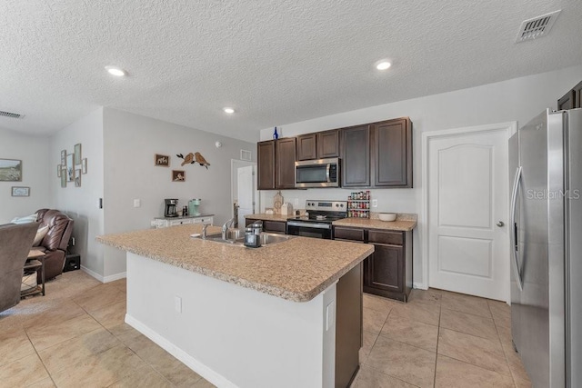 kitchen featuring a textured ceiling, light tile patterned flooring, an island with sink, appliances with stainless steel finishes, and dark brown cabinetry