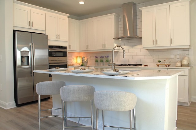 kitchen with stainless steel appliances, white cabinetry, a center island with sink, and wall chimney range hood