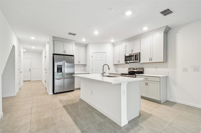 kitchen featuring stainless steel appliances, sink, gray cabinets, light tile patterned flooring, and a center island with sink