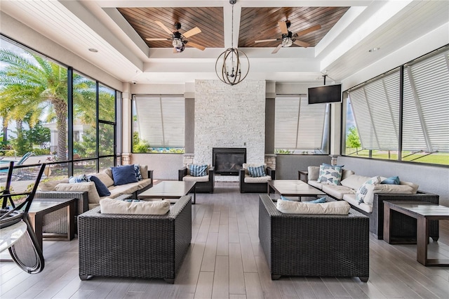 sunroom / solarium featuring ceiling fan with notable chandelier, a raised ceiling, wood ceiling, and a stone fireplace