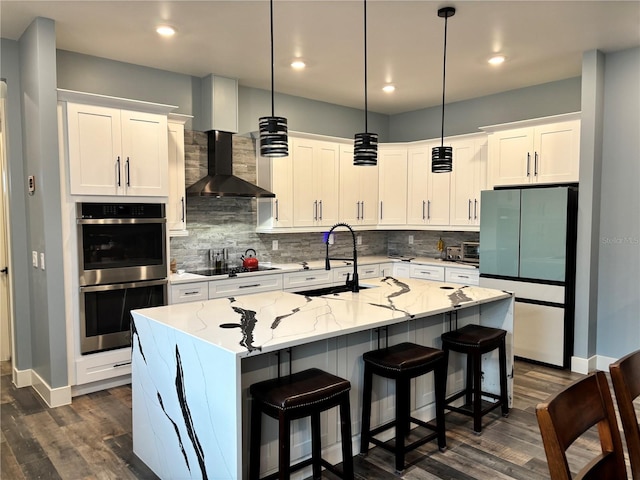 kitchen with decorative light fixtures, an island with sink, white cabinets, wall chimney exhaust hood, and light stone counters