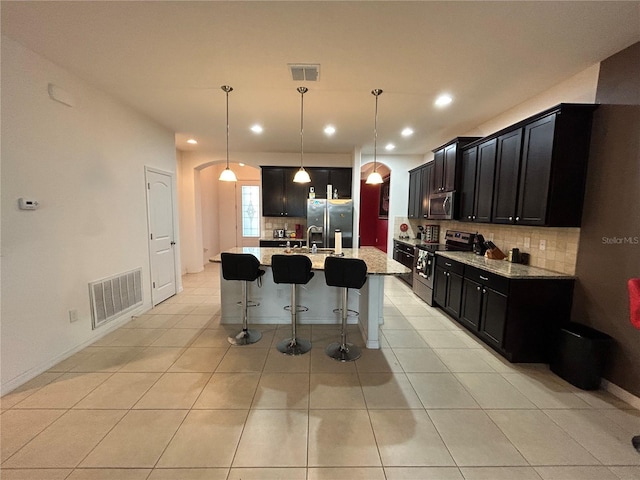 kitchen featuring pendant lighting, light tile patterned floors, appliances with stainless steel finishes, light stone countertops, and an island with sink