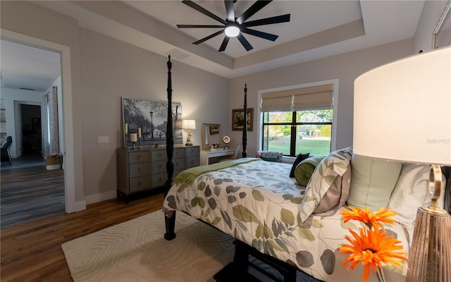 bedroom with ceiling fan, a tray ceiling, visible vents, baseboards, and wood finished floors