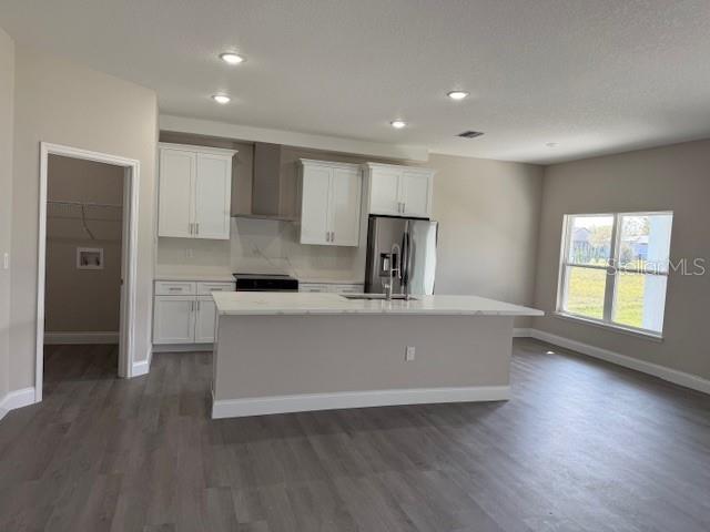 kitchen featuring an island with sink, stainless steel fridge with ice dispenser, a sink, white cabinets, and wall chimney range hood