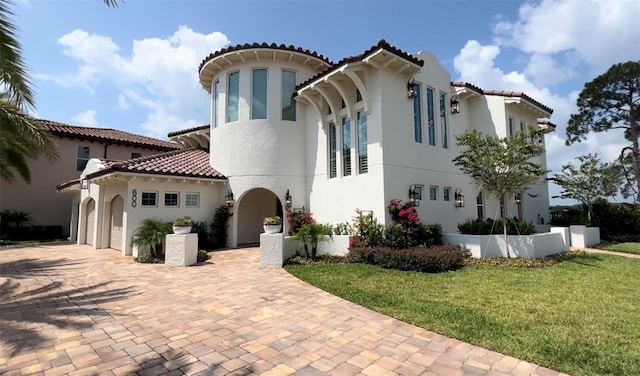 mediterranean / spanish-style home with a front lawn, a tiled roof, stucco siding, decorative driveway, and a garage