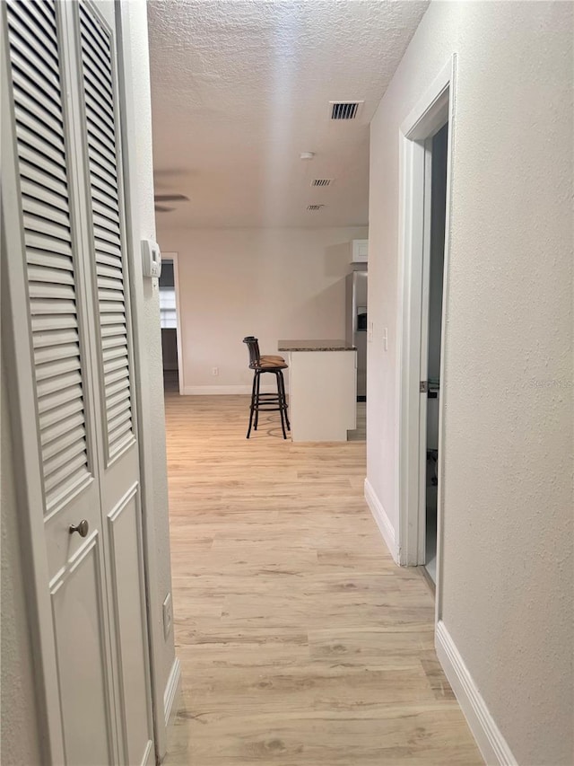 hall featuring a textured ceiling, visible vents, light wood-style flooring, baseboards, and a textured wall