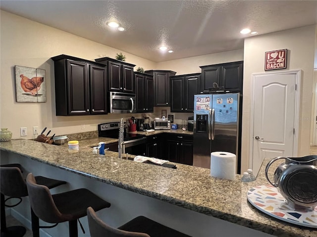 kitchen featuring appliances with stainless steel finishes, light stone counters, a textured ceiling, a kitchen bar, and kitchen peninsula