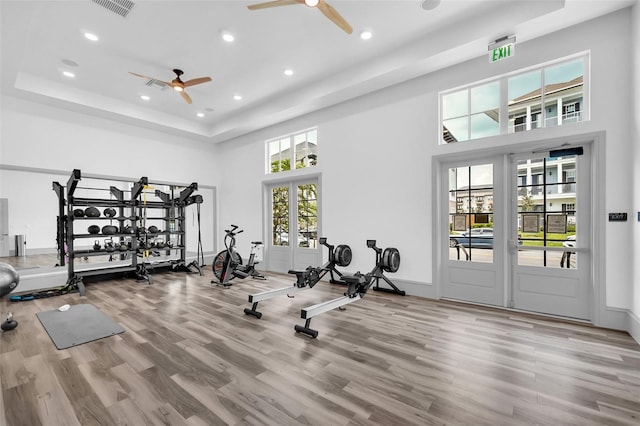 workout area with a towering ceiling, wood-type flooring, french doors, ceiling fan, and a tray ceiling