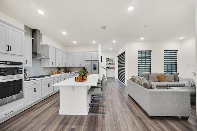 kitchen with white cabinets, wall chimney range hood, stainless steel appliances, a center island with sink, and a breakfast bar area