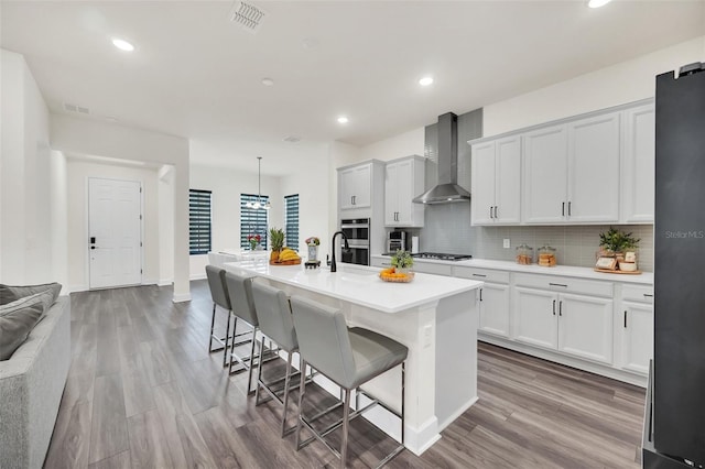 kitchen featuring wall chimney range hood, a breakfast bar, white cabinetry, hanging light fixtures, and a kitchen island with sink