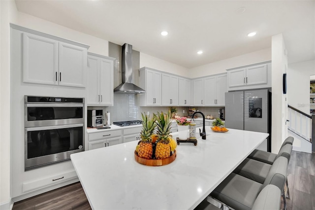 kitchen featuring appliances with stainless steel finishes, a kitchen bar, white cabinetry, wall chimney range hood, and a kitchen island with sink