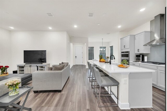 kitchen with light hardwood / wood-style flooring, a kitchen island with sink, a kitchen breakfast bar, stainless steel appliances, and wall chimney exhaust hood