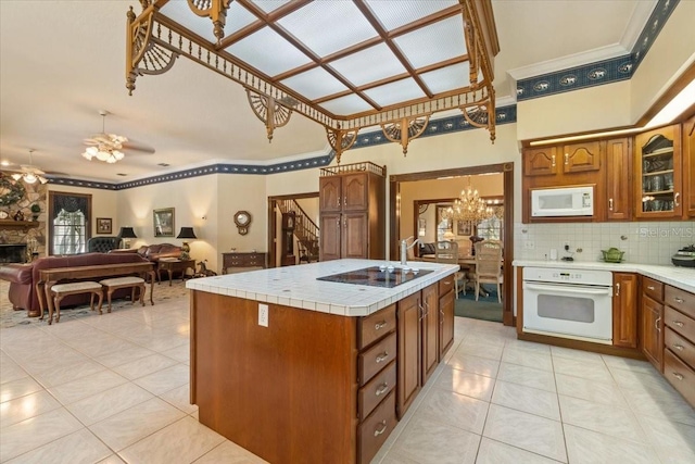 kitchen featuring ceiling fan with notable chandelier, tasteful backsplash, a center island with sink, a stone fireplace, and white appliances