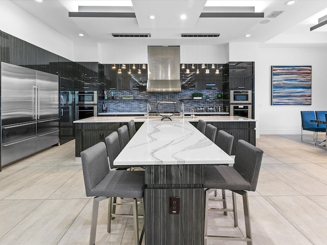 kitchen with pendant lighting, backsplash, a center island with sink, wall chimney exhaust hood, and a kitchen bar