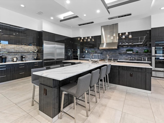 kitchen featuring an island with sink, stainless steel appliances, a kitchen bar, wall chimney exhaust hood, and light stone counters