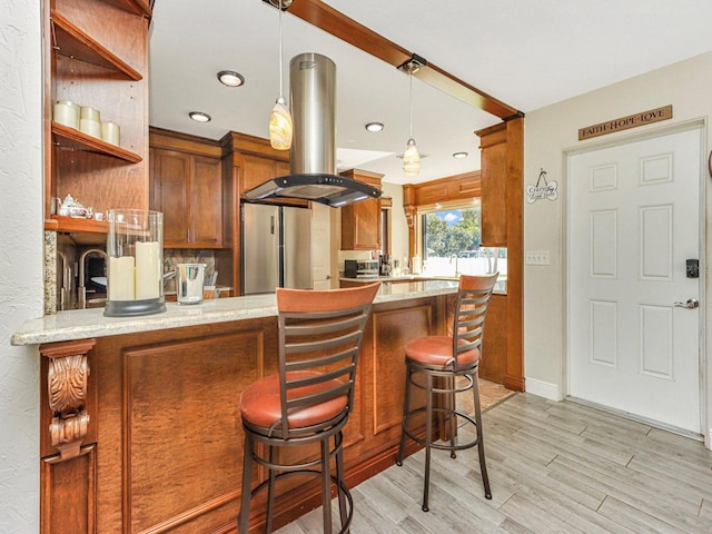 kitchen with stainless steel fridge, light stone counters, a breakfast bar area, island range hood, and decorative light fixtures