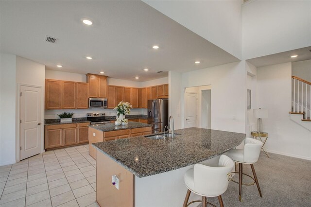 kitchen with a breakfast bar, dark stone counters, stainless steel appliances, light colored carpet, and a center island with sink