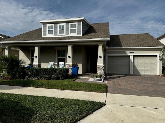 view of front of house with an attached garage, covered porch, a shingled roof, stucco siding, and decorative driveway