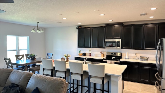 kitchen with stainless steel appliances, light tile floors, a kitchen bar, a kitchen island with sink, and an inviting chandelier