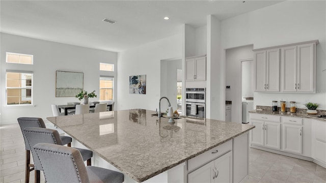 kitchen featuring double oven, an island with sink, light stone counters, and plenty of natural light