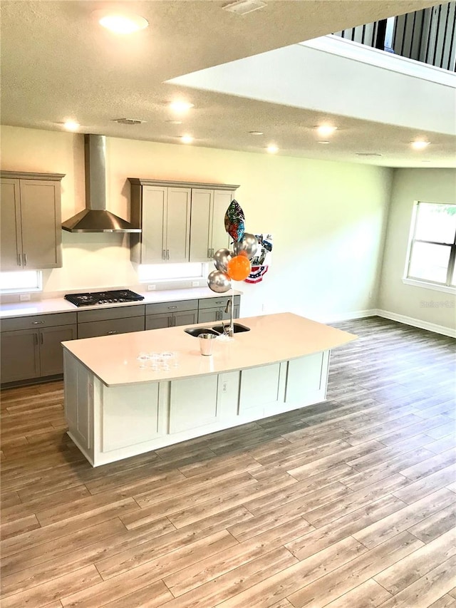 kitchen with light hardwood / wood-style flooring, wall chimney exhaust hood, black gas cooktop, and a kitchen island with sink