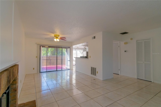 empty room with a textured ceiling, light tile patterned floors, a brick fireplace, and ceiling fan