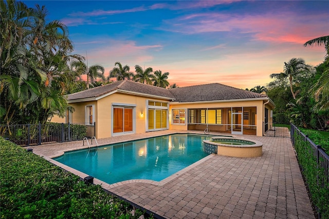 pool at dusk featuring a patio, a pool with connected hot tub, fence, and a sunroom