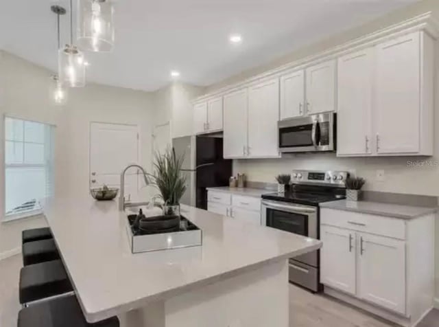 kitchen with an island with sink, stainless steel appliances, light countertops, white cabinetry, and pendant lighting