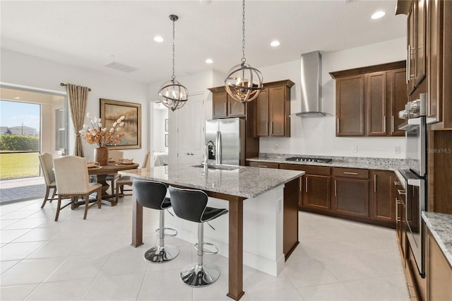 kitchen with light stone countertops, stainless steel appliances, wall chimney range hood, a kitchen island with sink, and pendant lighting