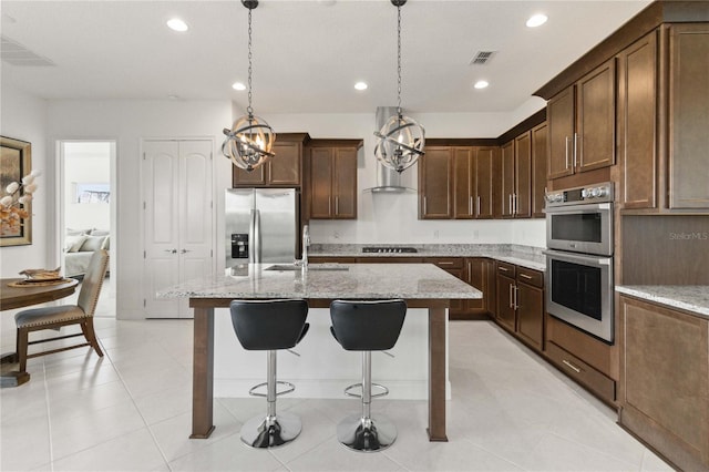 kitchen with light stone countertops, stainless steel appliances, a center island with sink, and decorative light fixtures