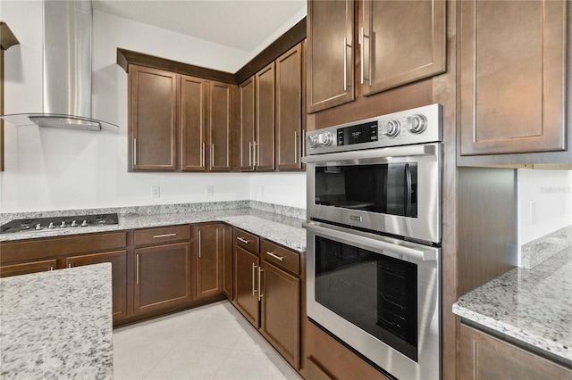 kitchen featuring wall chimney exhaust hood, light stone countertops, stainless steel appliances, light tile floors, and dark brown cabinetry