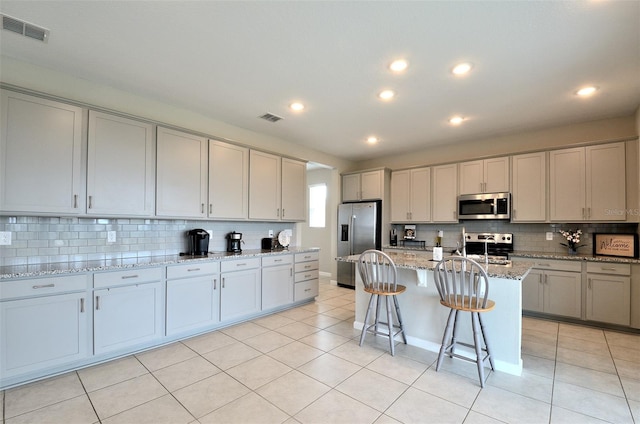 kitchen with backsplash, a center island with sink, light tile patterned flooring, light stone counters, and stainless steel appliances