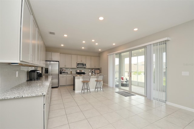 kitchen featuring backsplash, a center island with sink, light stone countertops, appliances with stainless steel finishes, and a kitchen bar