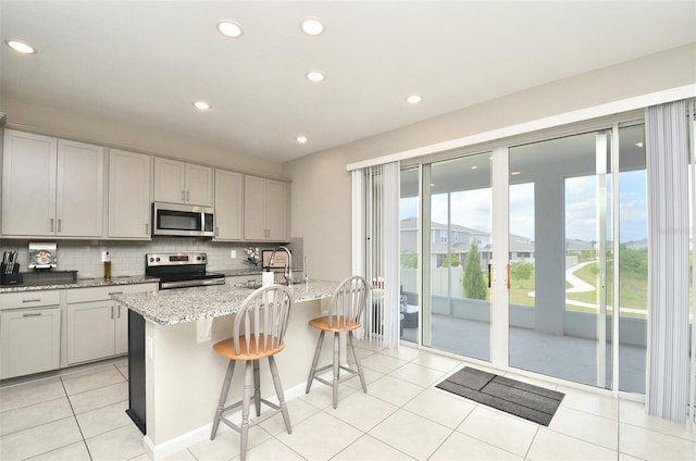 kitchen with backsplash, light stone countertops, an island with sink, appliances with stainless steel finishes, and a breakfast bar area