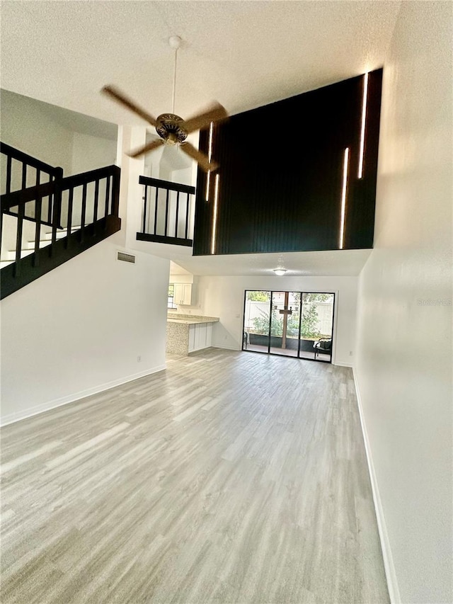 unfurnished living room with a textured ceiling, ceiling fan, a high ceiling, and light wood-type flooring