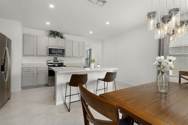 kitchen featuring gray cabinetry, stainless steel appliances, an island with sink, pendant lighting, and light tile flooring