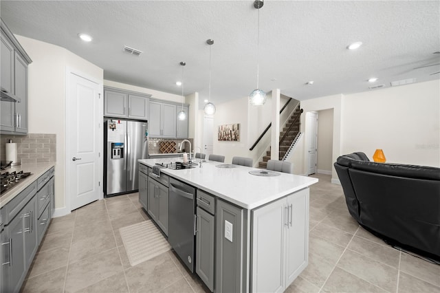 kitchen featuring stainless steel appliances, tasteful backsplash, a kitchen island with sink, gray cabinets, and pendant lighting