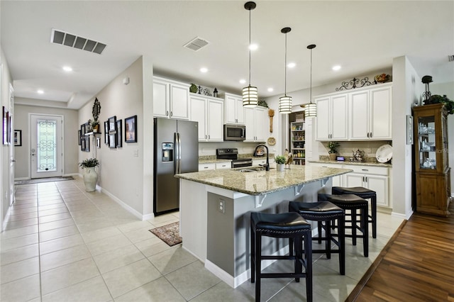 kitchen with white cabinetry, hanging light fixtures, stainless steel appliances, light stone counters, and a kitchen island with sink