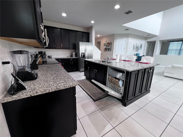 kitchen featuring decorative backsplash, stainless steel refrigerator with ice dispenser, light stone counters, sink, and an island with sink