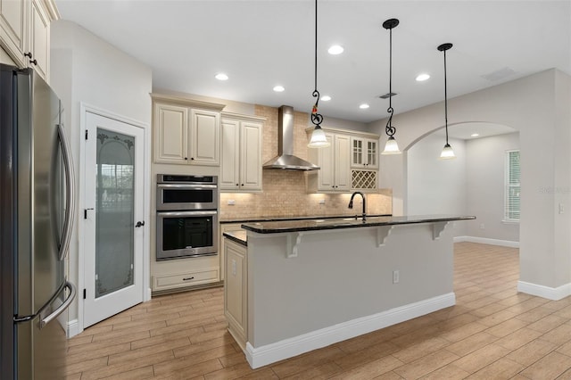 kitchen with wall chimney range hood, cream cabinetry, backsplash, a breakfast bar area, and appliances with stainless steel finishes