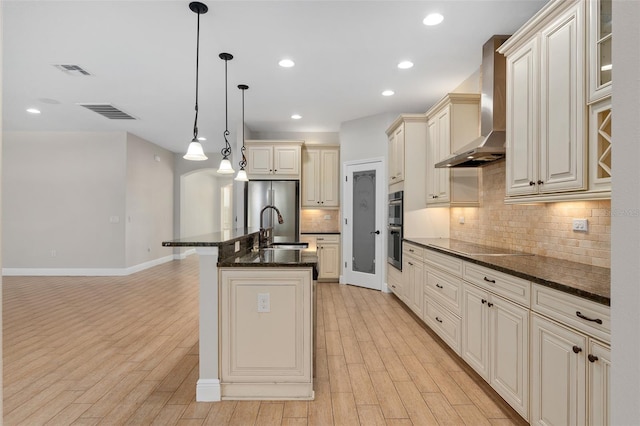 kitchen featuring wall chimney range hood, light hardwood / wood-style flooring, backsplash, hanging light fixtures, and a center island with sink