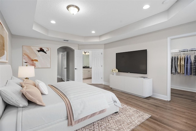 bedroom with a walk in closet, a tray ceiling, ensuite bath, wood-type flooring, and a closet