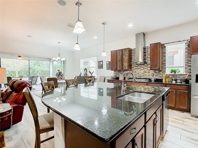 kitchen featuring an island with sink, sink, dark stone counters, hanging light fixtures, and wall chimney exhaust hood