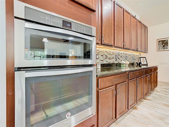 kitchen with stainless steel double oven, light wood-type flooring, decorative backsplash, and dark stone countertops