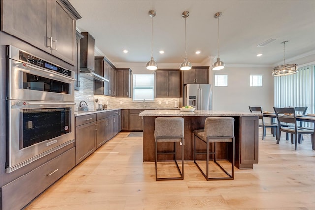 kitchen with wall chimney exhaust hood, ornamental molding, stainless steel appliances, decorative light fixtures, and a center island