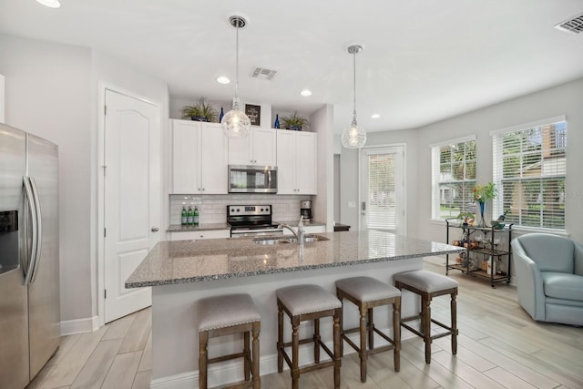 kitchen with a center island with sink, white cabinets, hanging light fixtures, light stone counters, and stainless steel appliances